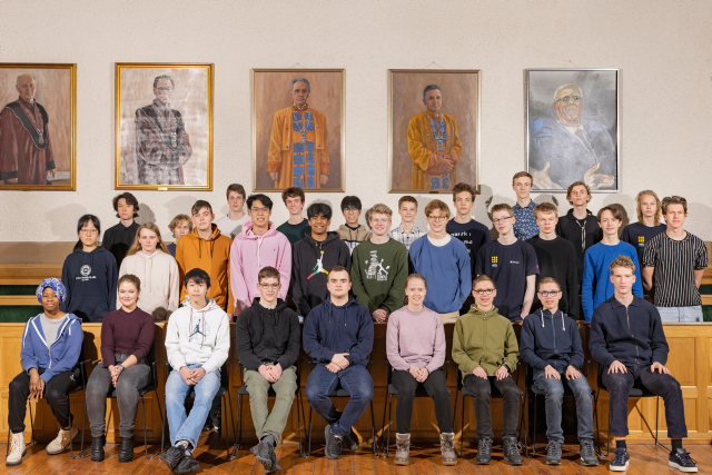 30 ungdommer oppstilt på et gruppebilde foran en vegg og malerier av eldre menn. 