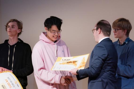 James Jia (18) får utdelt en premiesjekk fra statssekretær i kunnskapsdepartementet, Sindre Lysø. I bakgrunnen ser vi to gutter på ca. 18 år